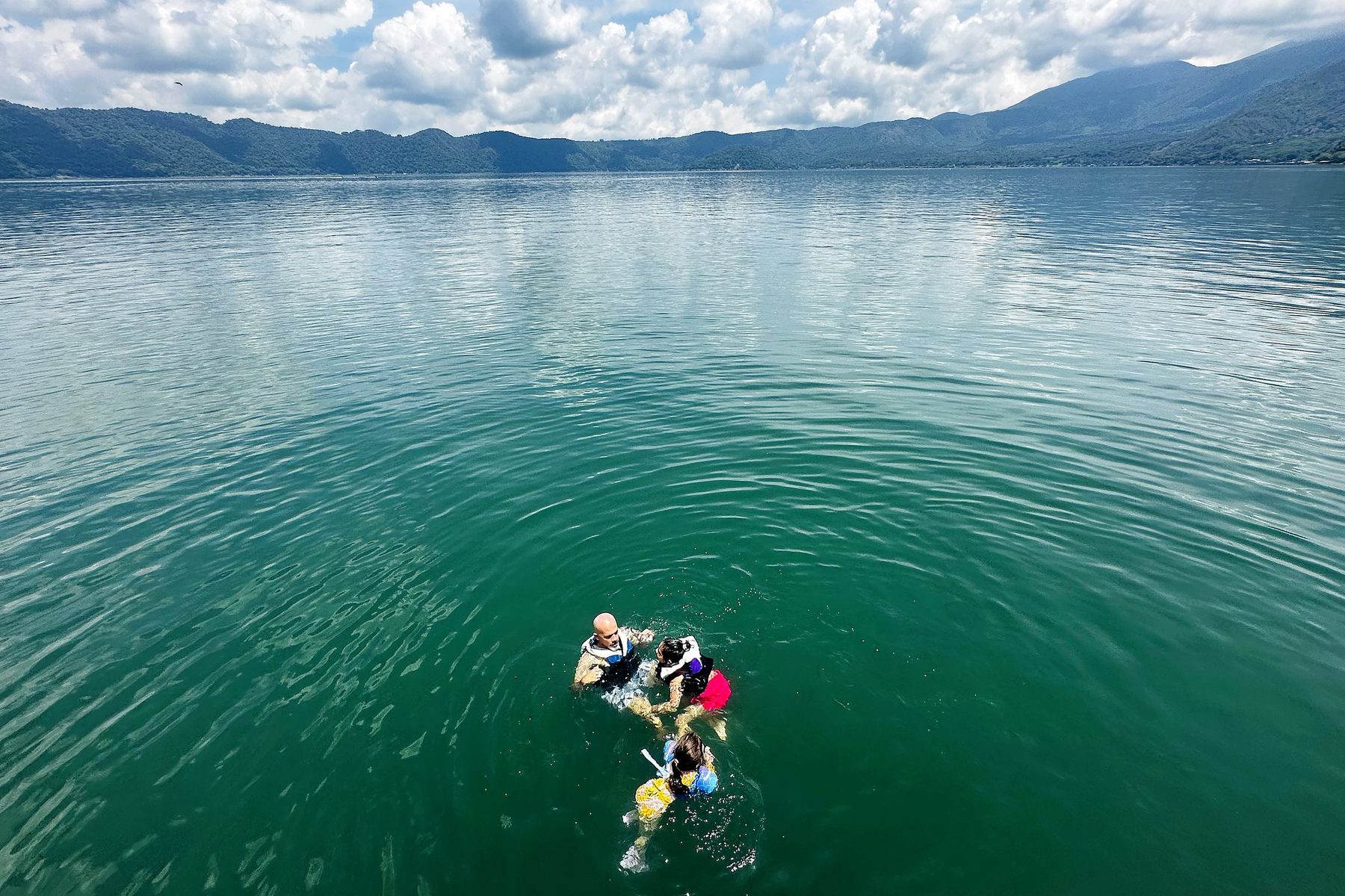 Our family swimming in life jackets in the green water of Lago de Coatepeque, with the crater rim and clouds behind